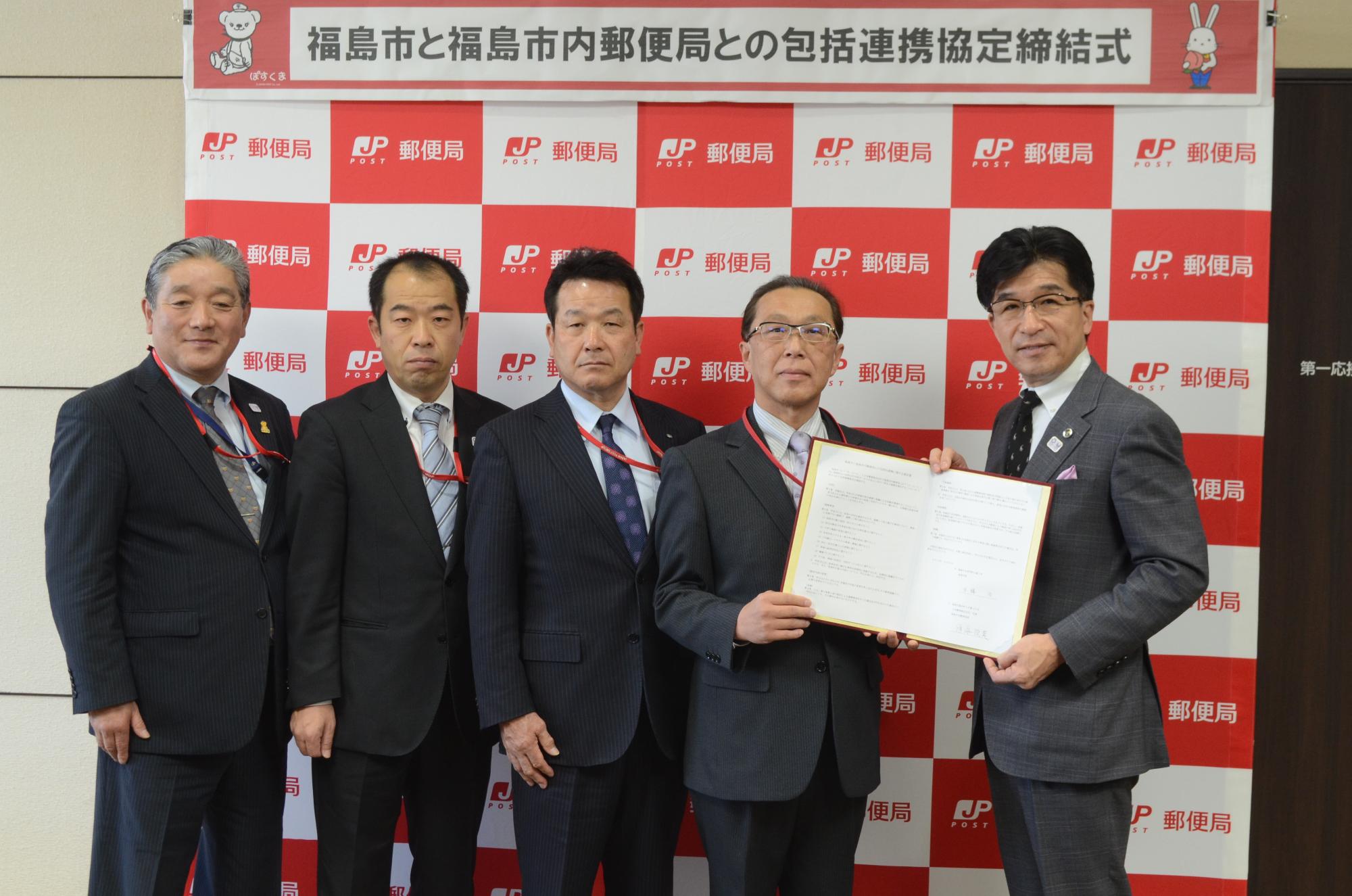 バックパネルの前で一緒に協定書を持つ市長と鳴海局長、3人の日本郵便株式会社の皆さんが横一列に並んでいる写真