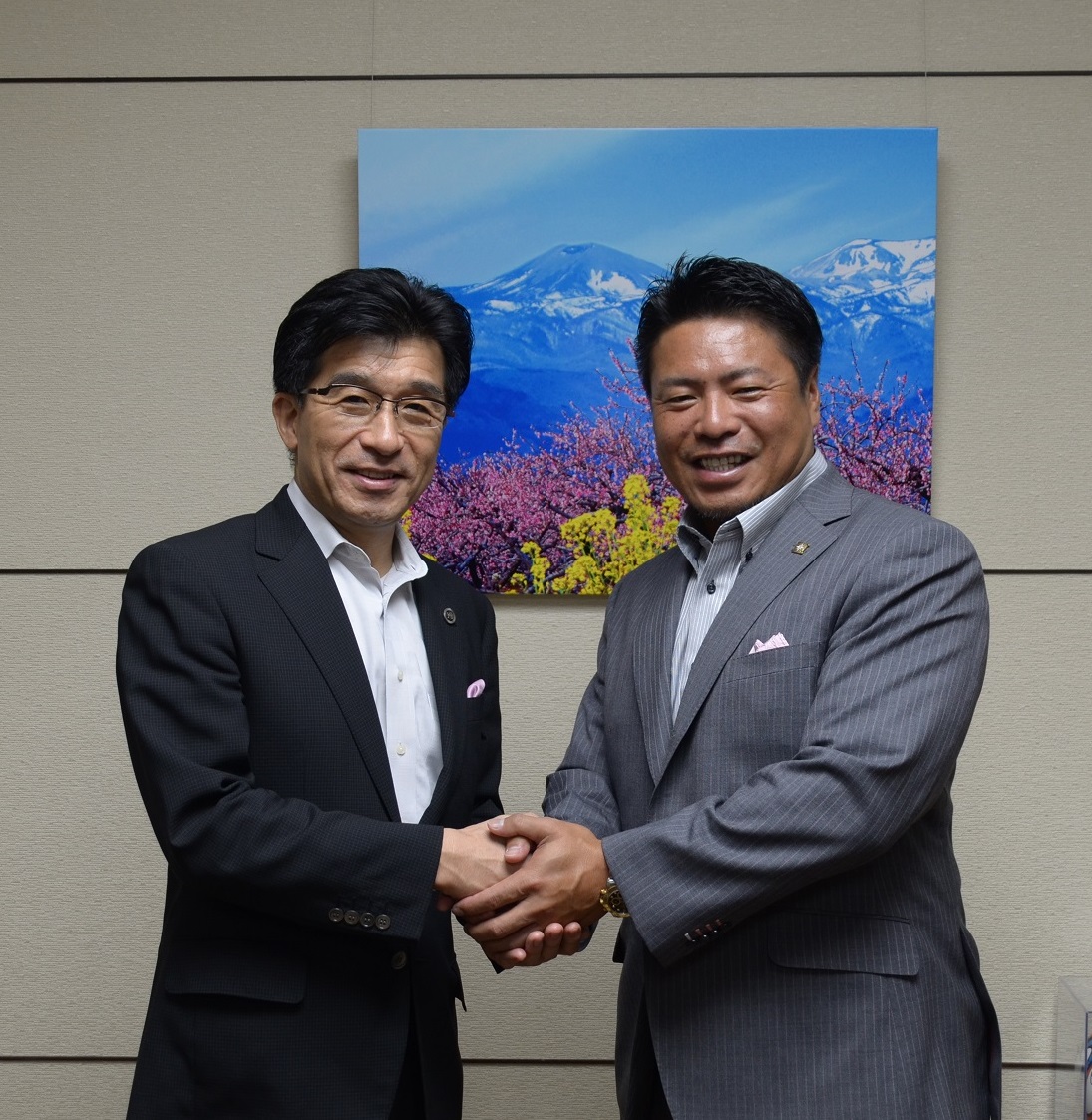 ピンク色と黄色の花と山の絵が飾られた室内で市長と岩村明憲監督が両手を握りあっている記念写真
