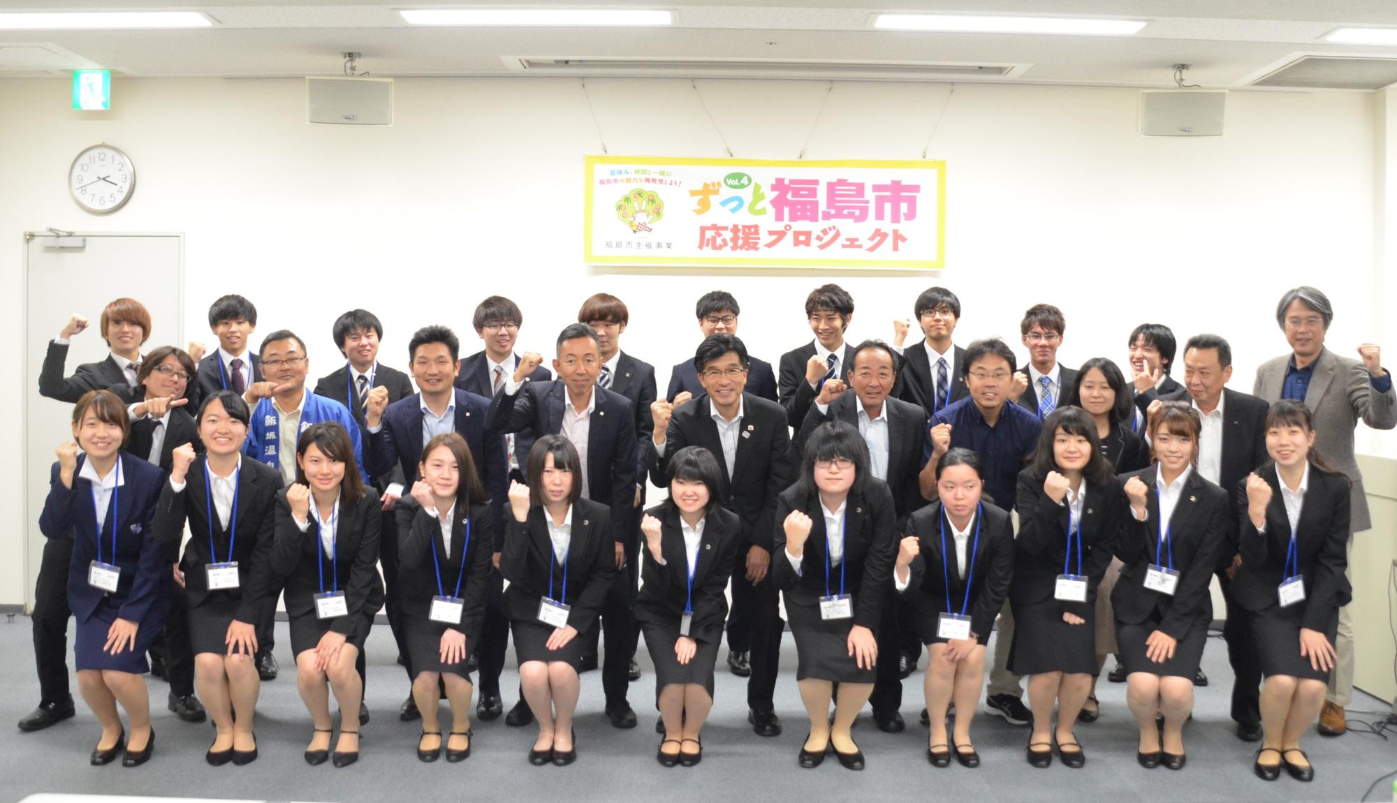 「ずっと福島市応援プロジェクト」の吊りパネルの前で、市長とスーツ姿の学生たち、関係者が3列に並び握りこぶしでポーズを取っている写真
