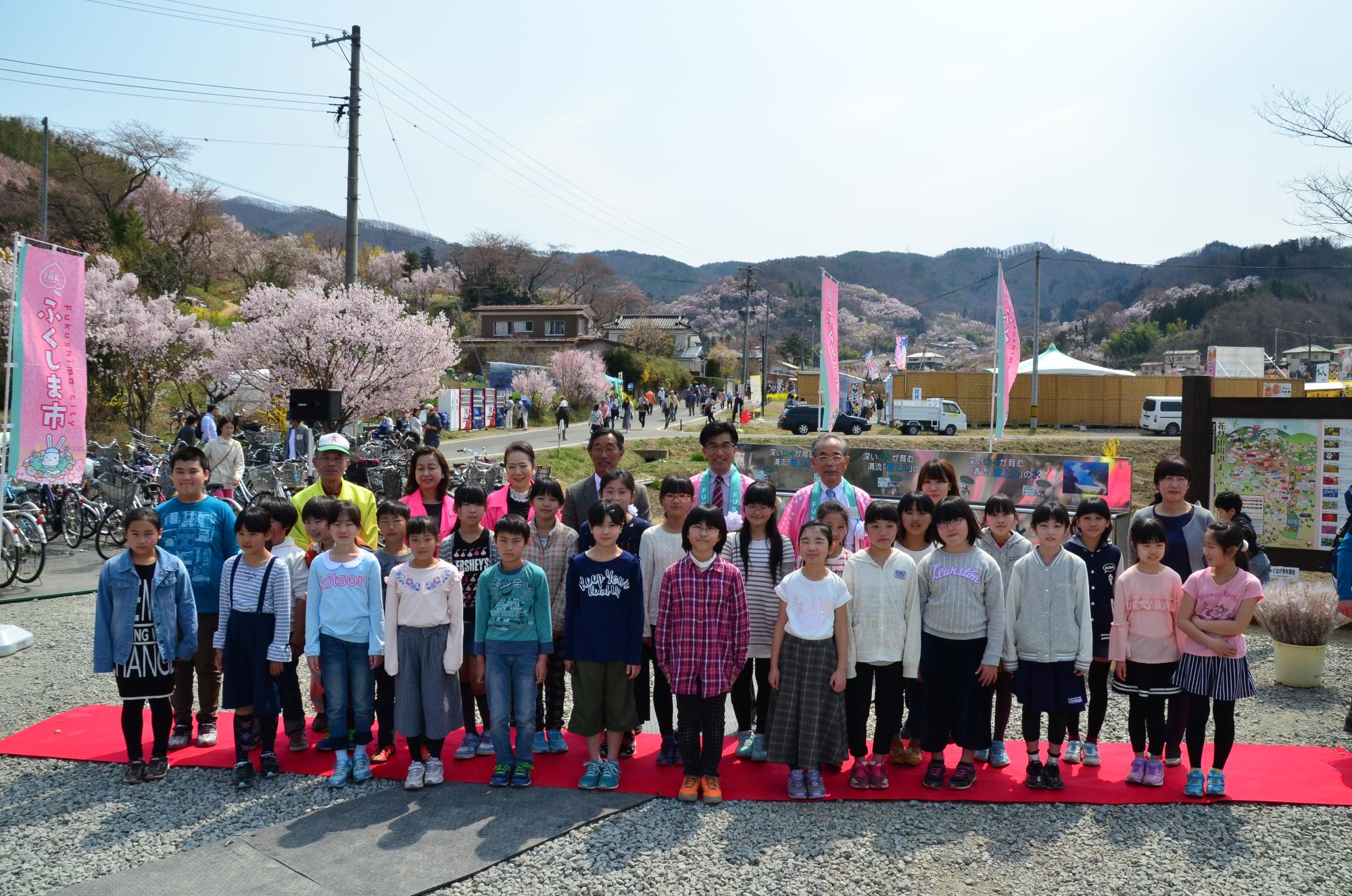 桜が咲いた広場に敷かれた赤いカーペットに、子供たちと関係者が横3列に並んだ集合写真
