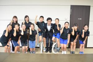 黒いTシャツに短パン姿の小学生達と関係者が握りこぶしでポーズを取っており、中央に一輪車にまたがる小学生を手で支えている市長の写真