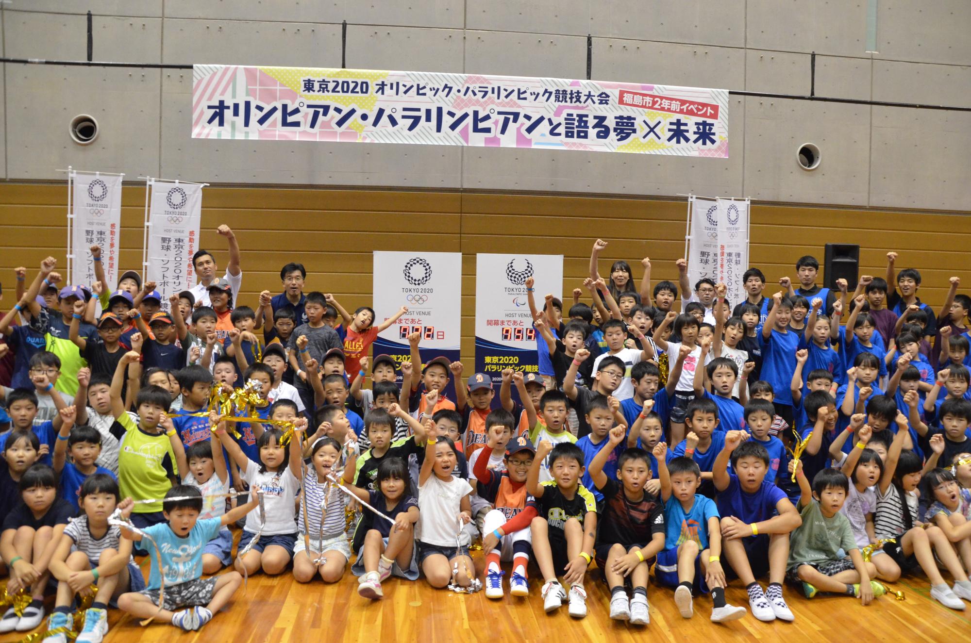 「オリンピアン・パラ臨費案と語る夢×未来」の横断幕があり、市長と小学生くらいの子供達、関係者が集合し、握りこぶしでポーズを取っている写真