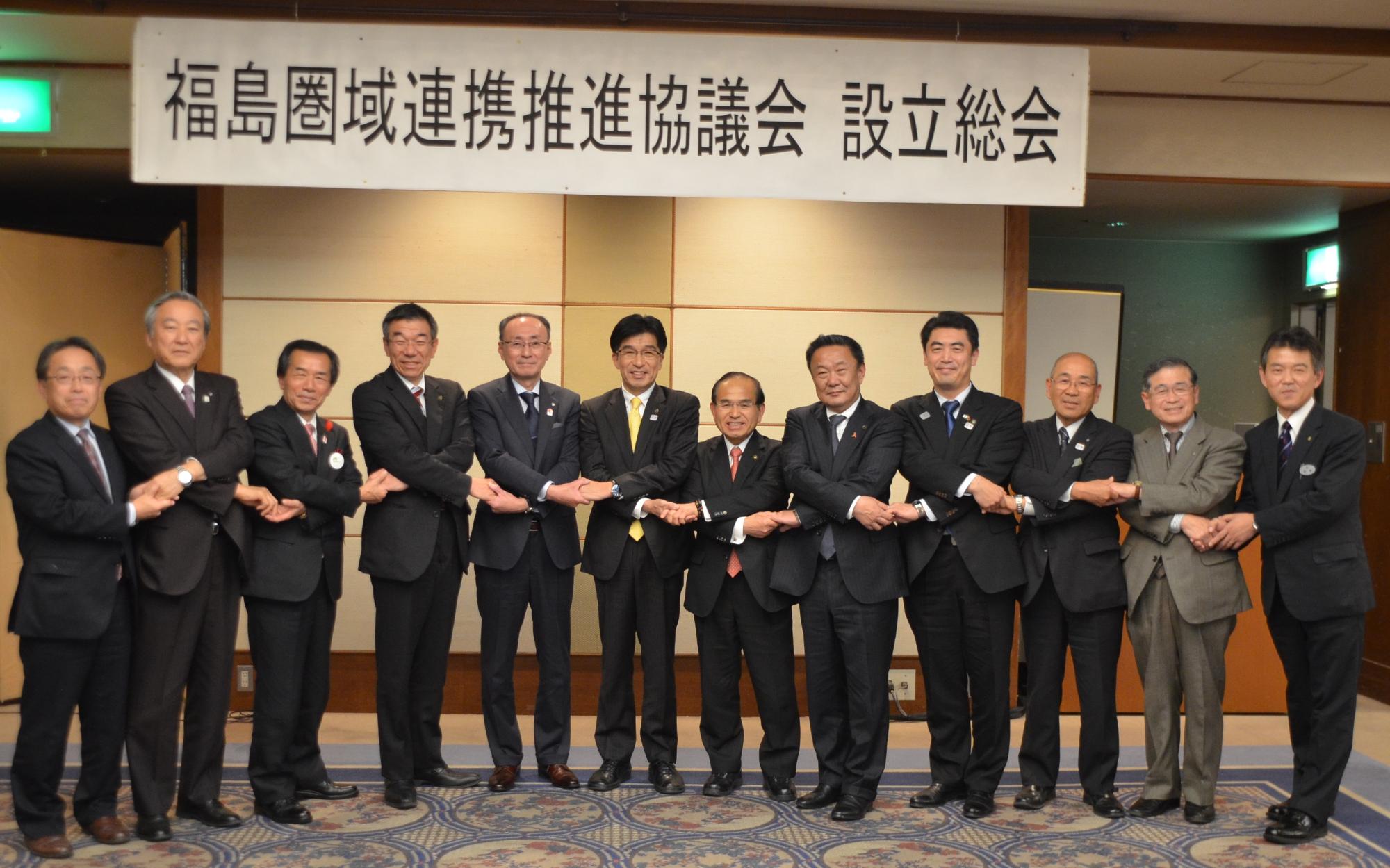 「福島圏域連携推進協議会設立総会」の吊りパネルの下で、市長と11市町村長が両手を前でクロスして隣の人と手を組んでいる写真