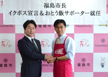 「福島市長イクボス宣言&おとう飯サポーター就任」と張られた福島市のバックパネル前で、エプロン姿の市長と関係者が握手を交わしている写真