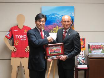ユニフォームやボール、選手たちの写真などが並べられており、市長と福島ユナイテッドFCの田坂監督が、額縁に入った選手の集合写真を手に持ちながら握手を交わしている写真
