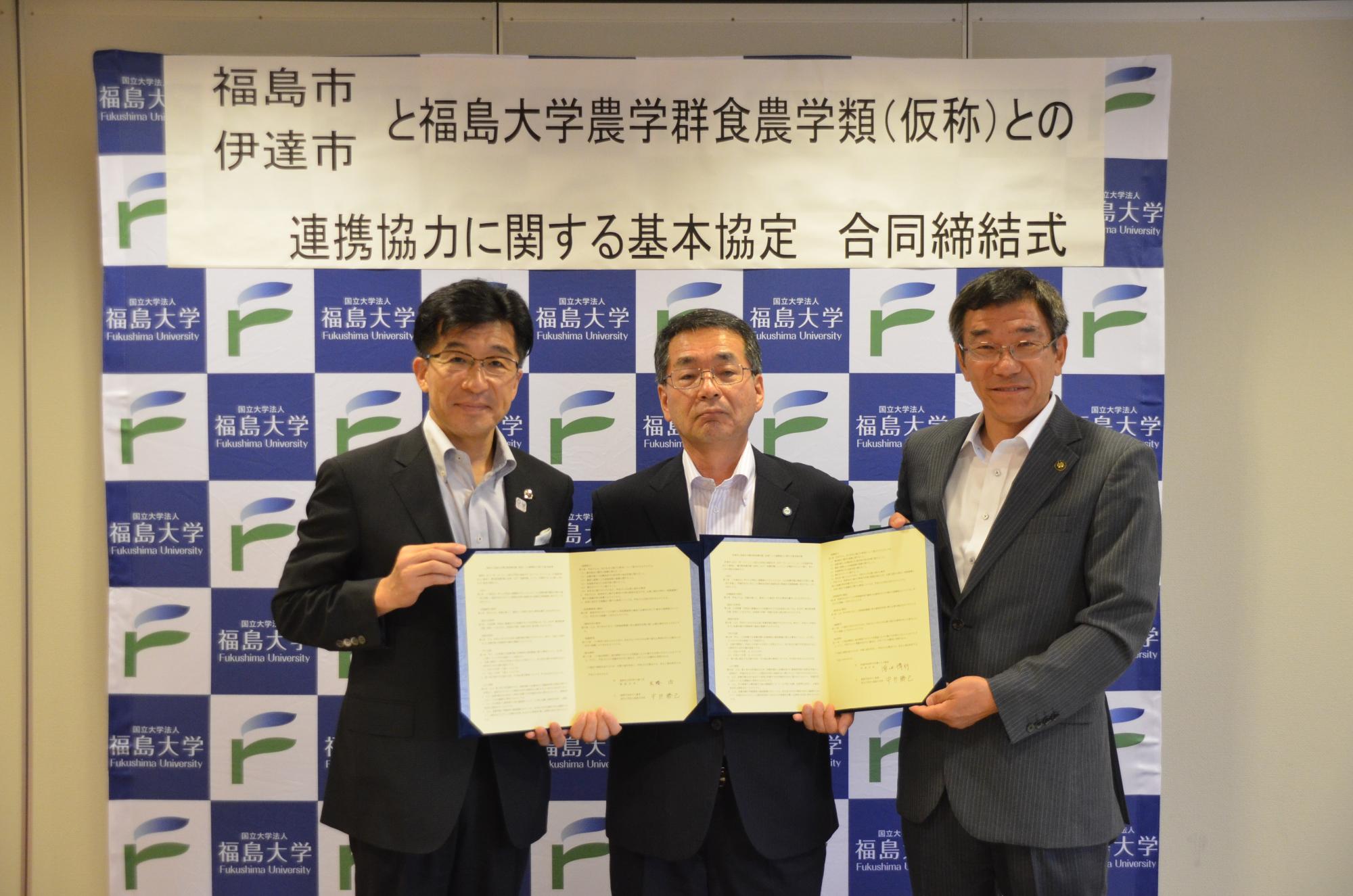 「福島市・伊達市と福島大学農学群食農学類(仮称)との連携協力に関する基本協定合同締結式」と書かれた福島大学のバックパネル前で、市長と関係者が締結書を開いて手に持っている写真
