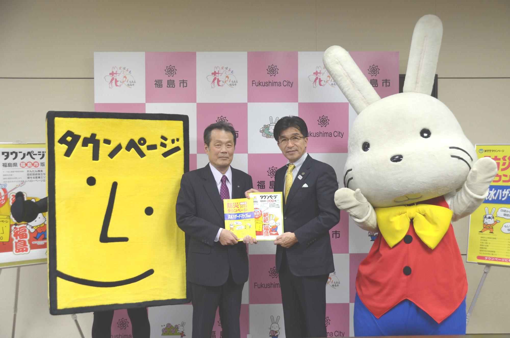 福島市のバックパネル前で、タウンページと書かれた黄色い四角のマスコットとももりんとの間に、市長と吉松常務が防災タウンページを手に持っている写真