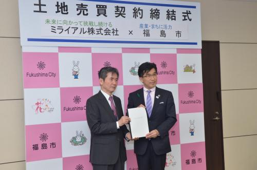 「土地売買契約締結式」と書かれた横断幕が掲げられた福島市のバックパネルの前で、ミライアルの代表の男性と市長が書類に手を携えて立っている写真