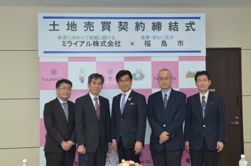 「土地売買契約締結式」と書かれた横断幕が掲げられた福島市のバックパネルの前に市長を中央に5人の男性が並んで立って写した記念写真