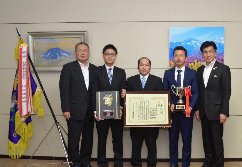 山の絵などが飾られた壁の前で、左に置かれた優勝旗の横に盾、賞状、優勝杯を胸の前で持った4名のスーツ姿の男性と市長が並んでいる記念写真