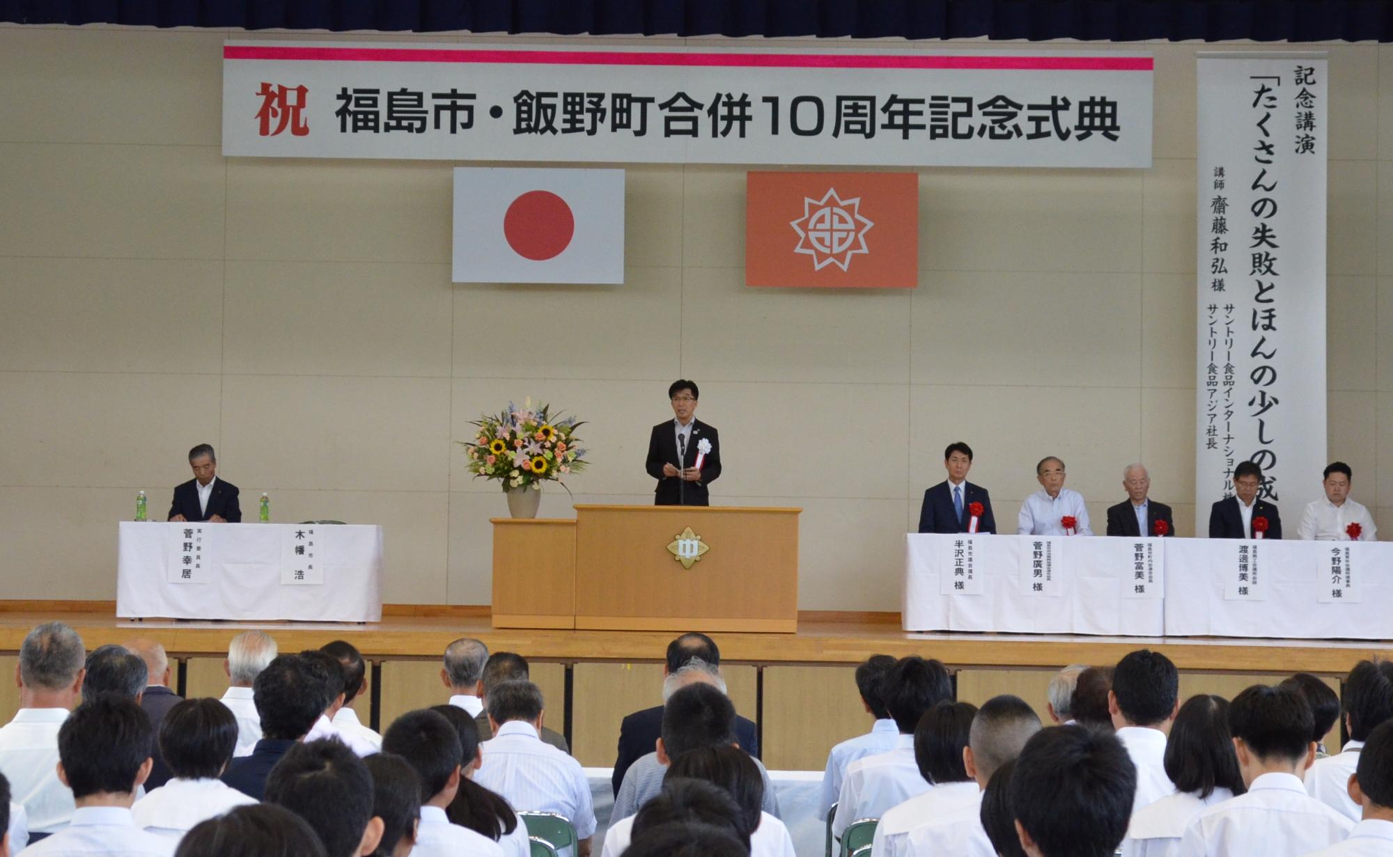 「福島市・飯野町合併10周年記念式典」の吊りパネルの下、来賓席に関係者が座っており、舞台の演壇で挨拶をする市長と、話に耳を傾ける参加者の写真