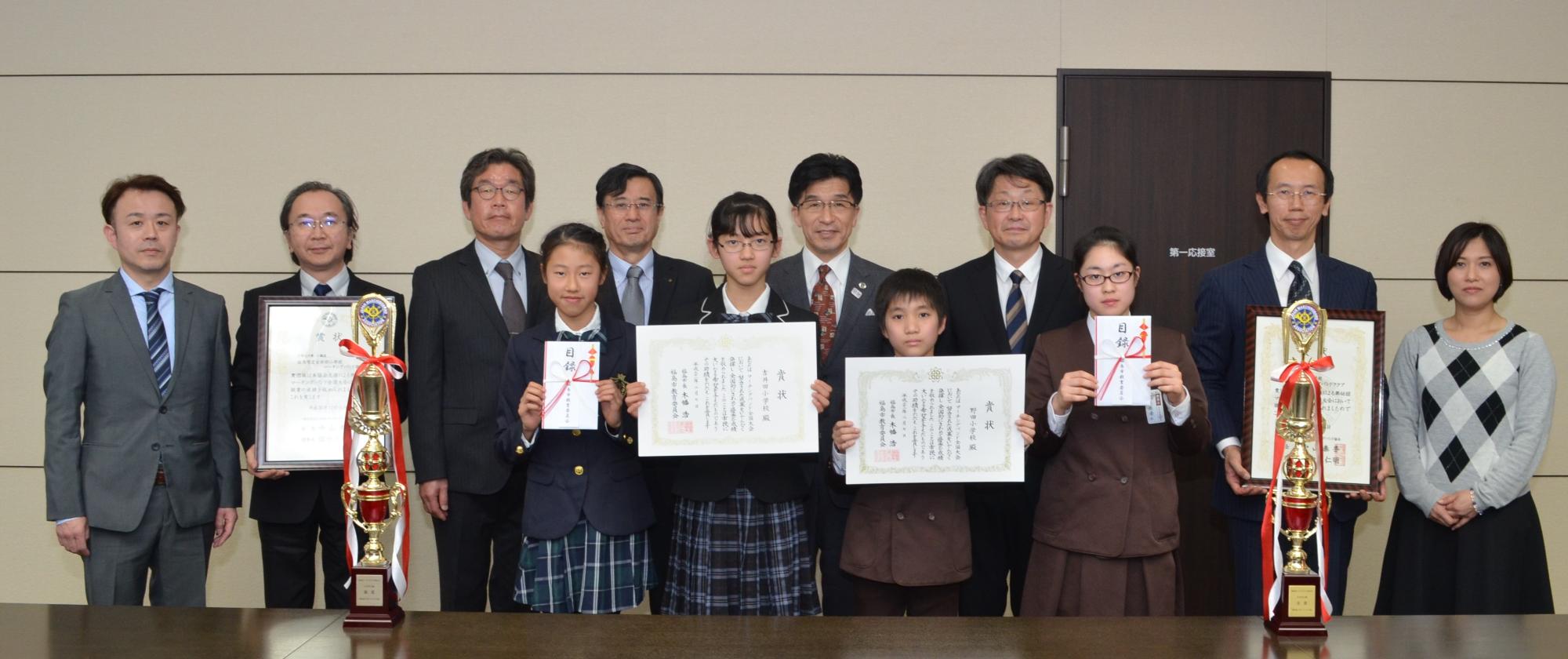 机の上にトロフィーが2つ置いてあり、制服姿の野田小学校・吉井田小学校の皆さんが目録と賞状を手に持ち、関係者らが並んでいる写真