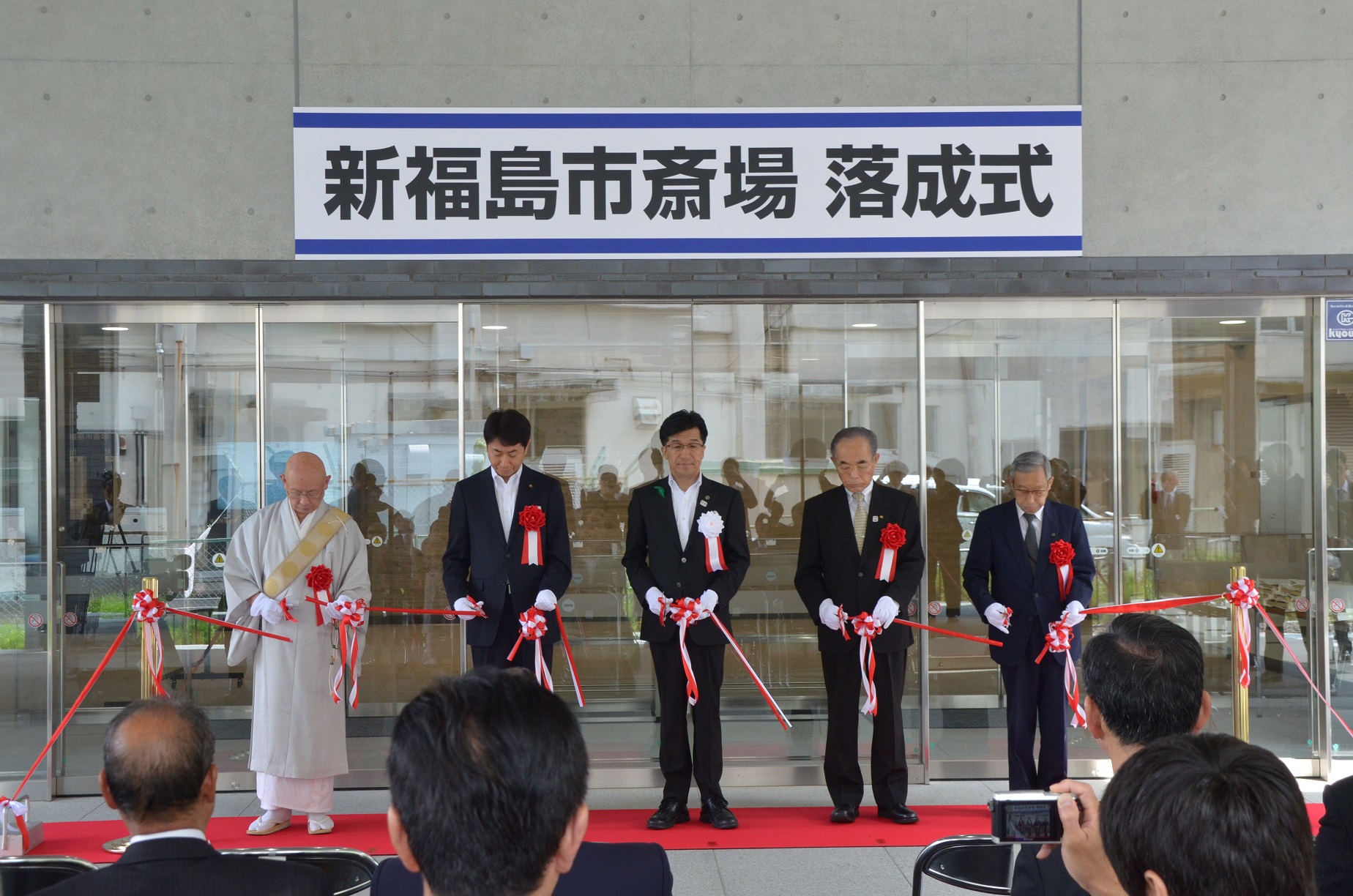 「新福島市斎場落成式」と書かれた吊り看板パネルの下にレッドカーペットが敷かれ、僧侶様とスーツを着た男性が並びテープカットをしている写真