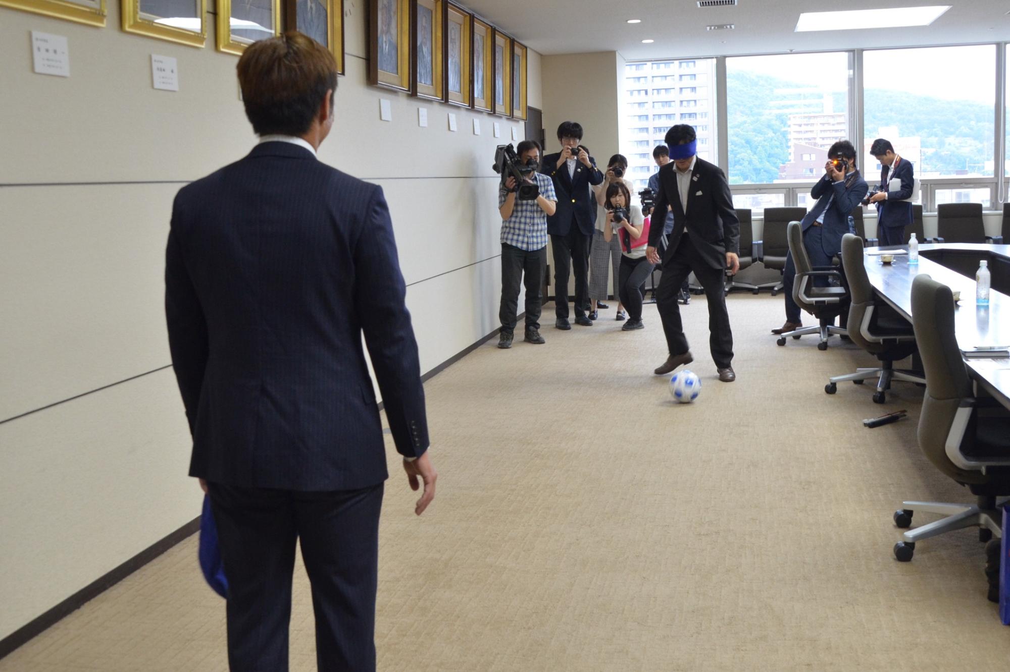 市長と加藤健人選手が室内でサッカーのパスをしているところをカメラマンが撮っている写真