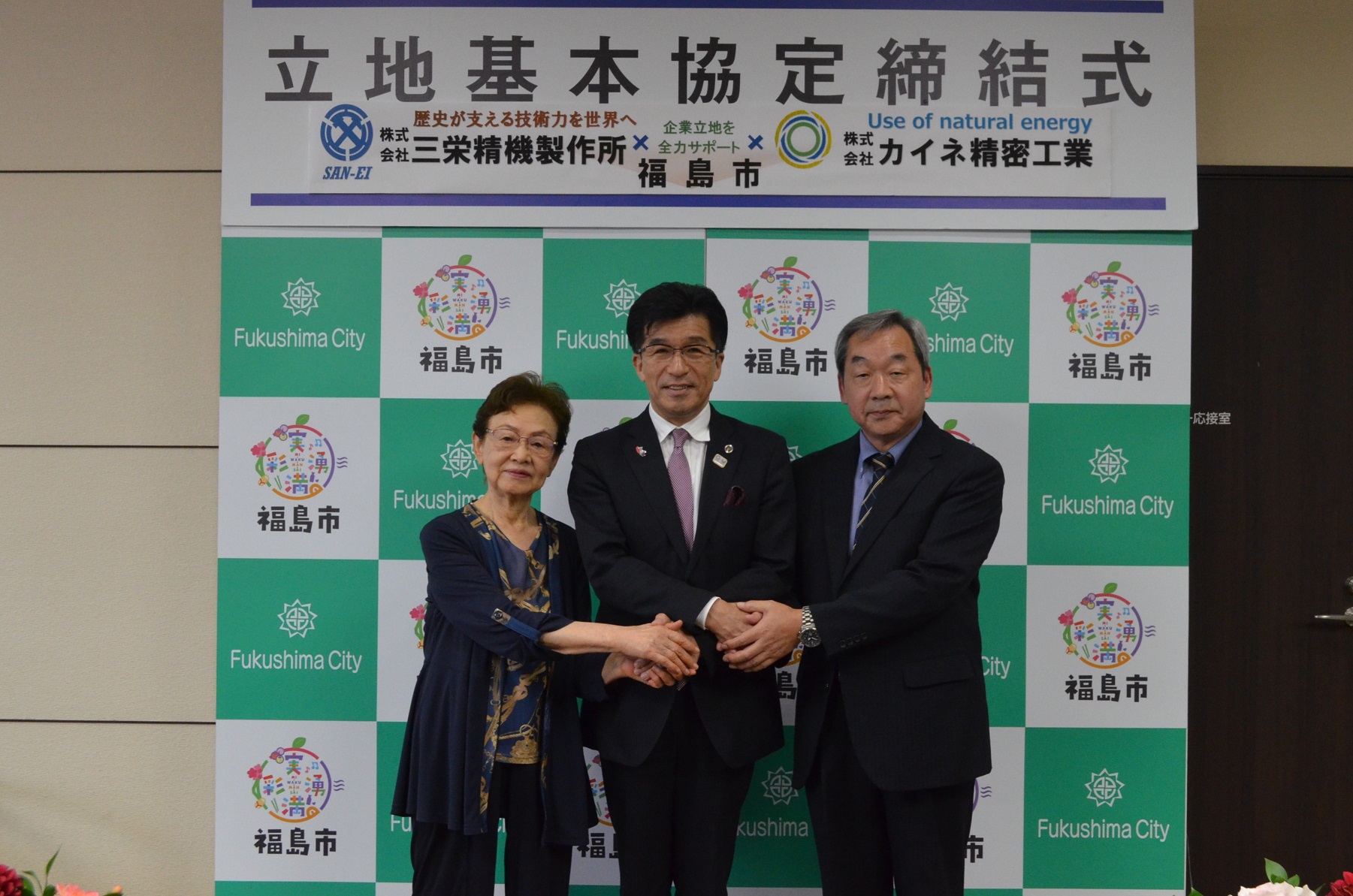 「立地基本協定締結式」と書かれたパネルの下に設置された福島市のバックボードの前で市長と株式会社三栄精機製作所の代表と株式会社カイネ精密工業の代表がASEAN式握手をしている写真