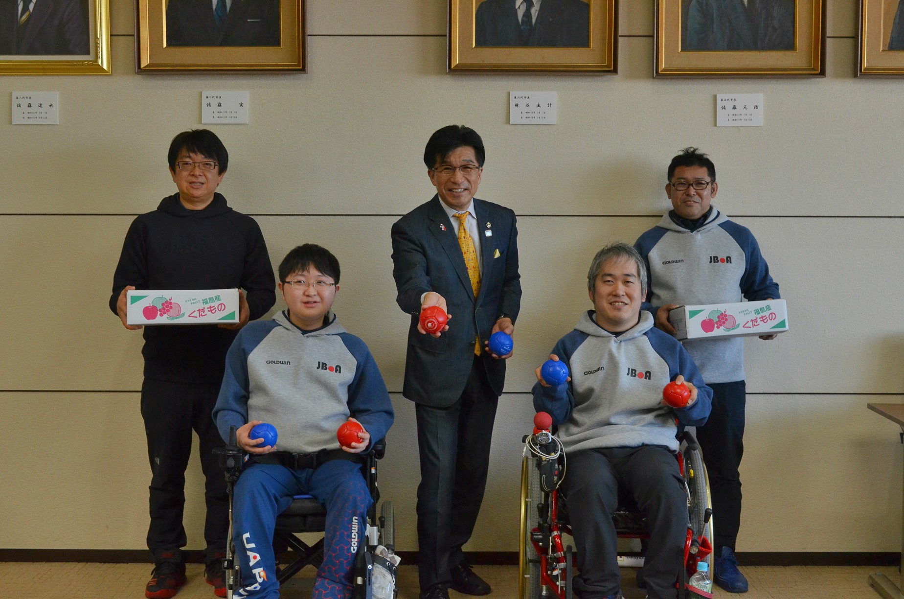 市長と市長の両隣りに車いすの乗った男性が手のひらサイズの赤いボールと青いボールを持っており、両脇に果物ダンボールを持った男性2人が立って記念撮影している写真