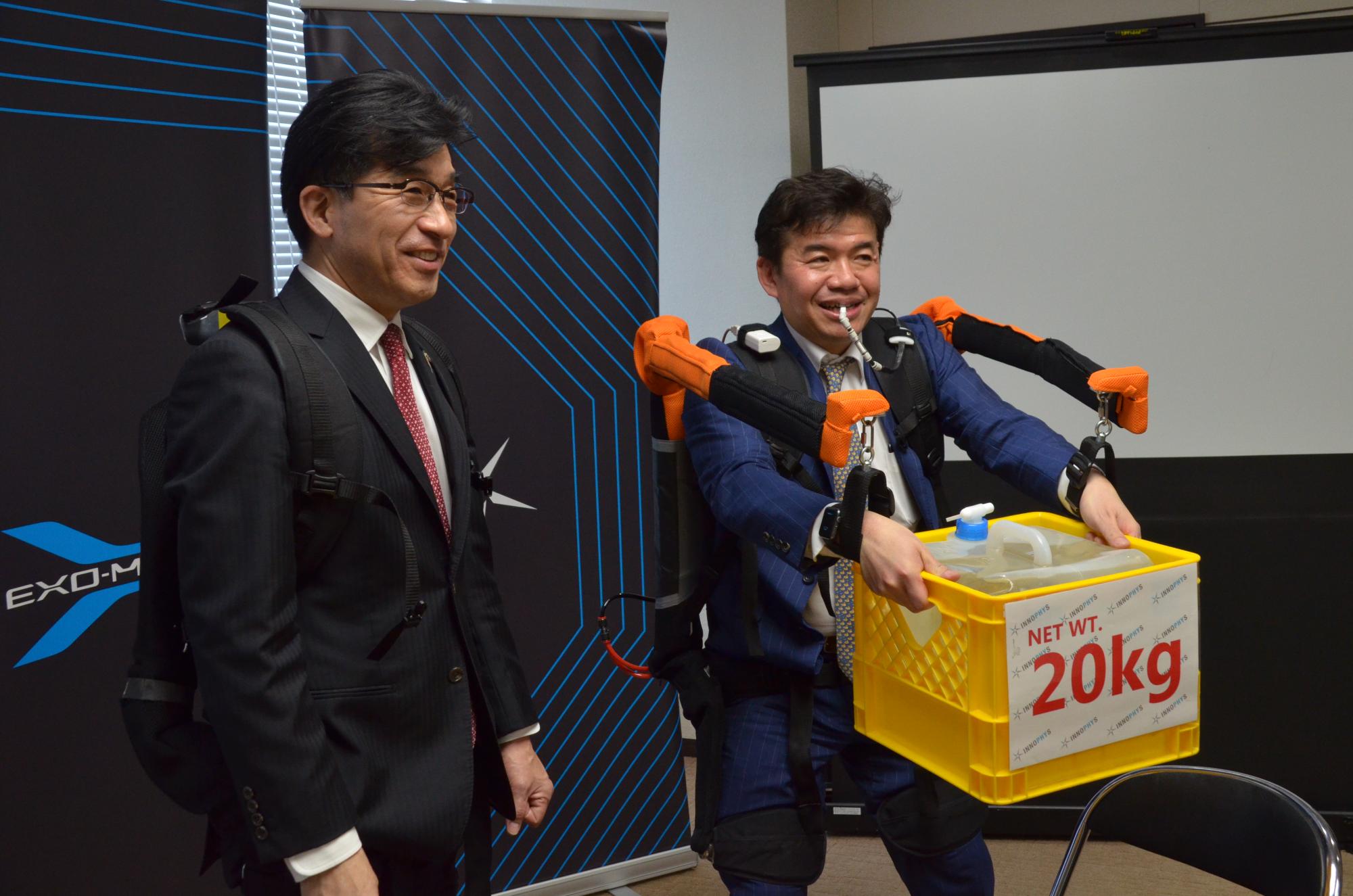 笑顔で立つ市長の隣で装着型アシストスーツを試用した古川社長が、赤文字で「20キログラム」と書かれた紙が貼られた黄色のカゴを前にして持っている様子の写真