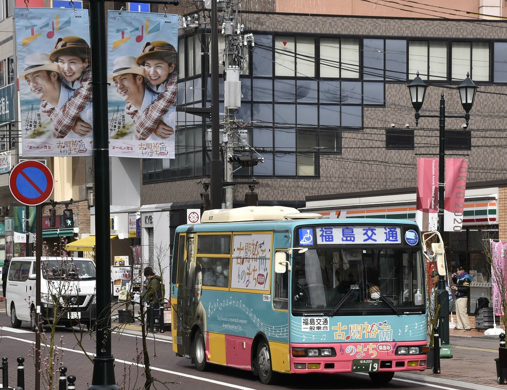 連続テレビ小説「エール」の袖型広告がある市道を水色やピンクや黄色で彩られたラッピングバスが走っている写真