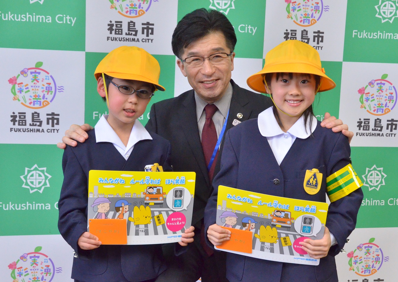 福島市のバックボードの前で黄色の帽子をかぶった男子児童と女子児童が交通安全用具を両手に持ち、児童の肩に手を添えた市長がかがみ記念撮影をしている写真