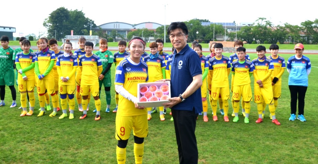 芝生の上で黄色のユニホームを着たベトナム女子サッカー代表が並び、代表で1人の選手にモモを贈呈する市長の写真