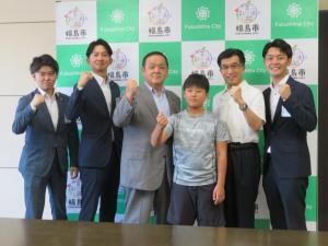 バックパネルの前で、男の子と市長を含めた男性5名が立ちガッツポーズをとっている写真