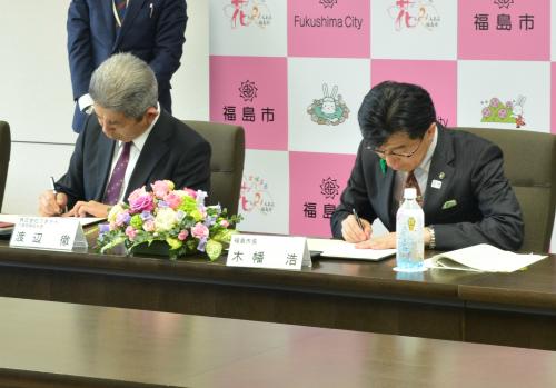 福島市のバックパネルの前に置かれたテーブルの席に着いた代表の男性と市長が書類にサインを行っている様子の写真