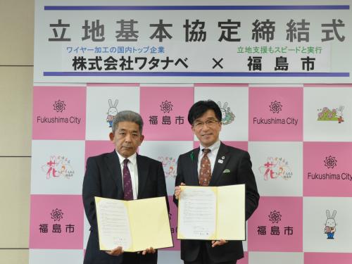 「立地基本協定締結式」と書かれた横断幕が掲げられた福島市のバックパネルの前で、代表の男性と市長が書類を開いて持ち並んで立っている写真