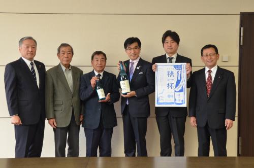 6人の男性が並んで立ち、中央の代表の男性と市長が緑色の瓶その右隣の男性が「精一杯」の文字と日本酒の瓶が青で書かれた紙を持っている写真