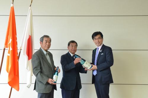 左側に赤い旗と国旗が並べて掲げられており、その横で緑色の瓶を持った代表の2人の男性と市長が並んで立っている写真