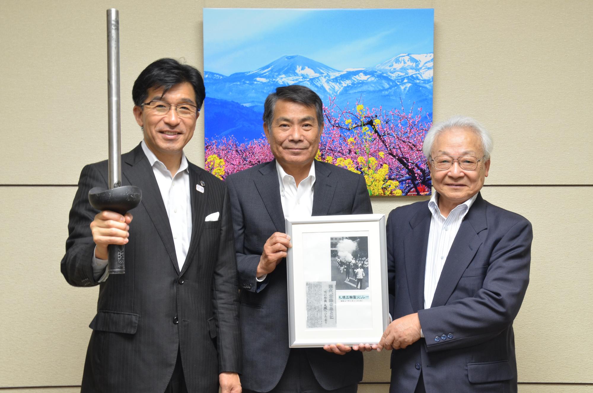 市長が札幌オリンピック聖火トーチを手に持ち、関係者が新聞記事の切り抜きが収められた額縁を手に持っている写真