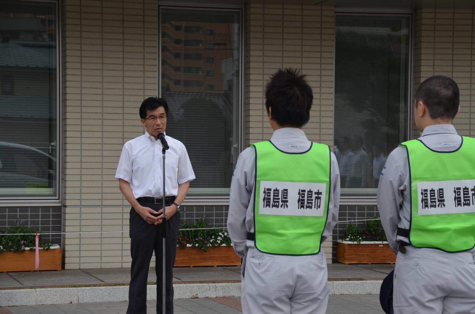 屋外で、グレーの作業服の上に蛍光グリーンで中央に「福島県福島市」と印刷されたビブスを着た男性2名と向かい合って立った市長がスタンドマイクの前で話をしている写真