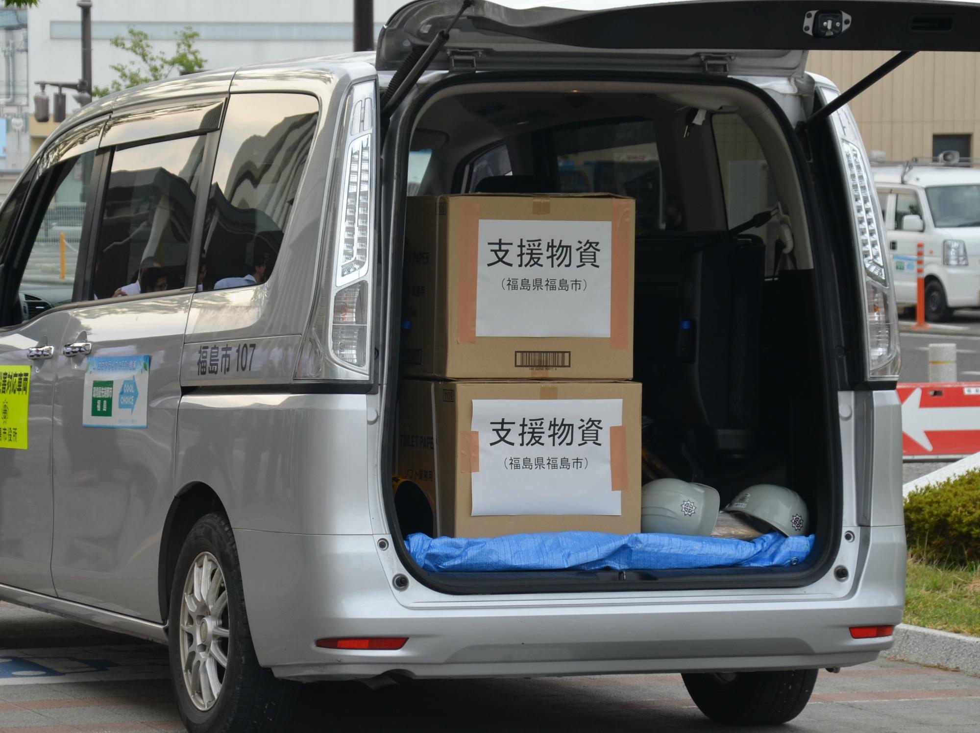 シルバーのバンのバックドアが開いており、車の中に支援物資と書かれた紙が貼りつけられた大きな段ボール箱が2段に重ねて積まれている様子の写真