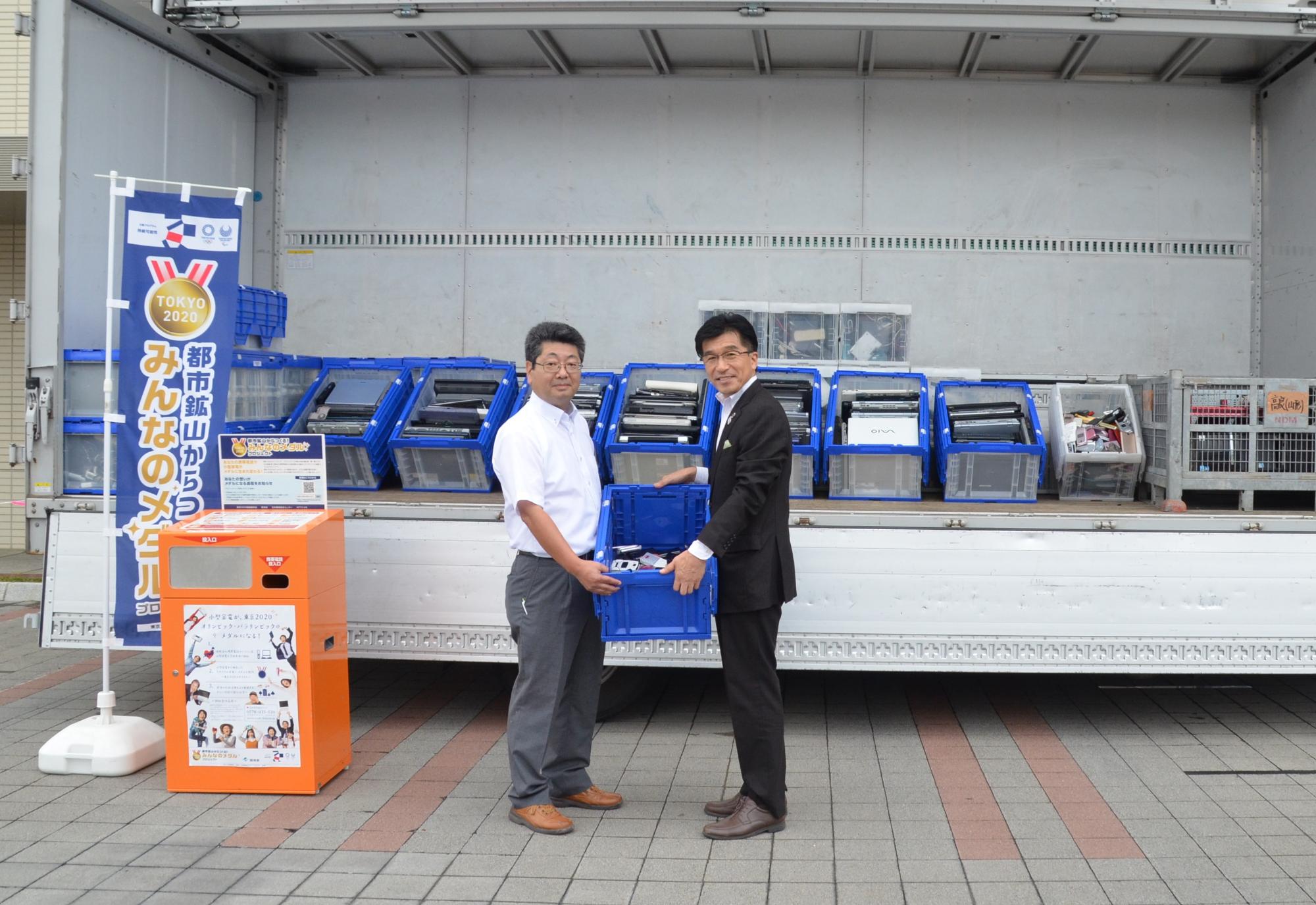 ウィング車に、パソコンや携帯電話などの小型家電がプラスチックの箱が積まれており、その前で市長と関係者が小型家電の入った箱を両手に持っている写真