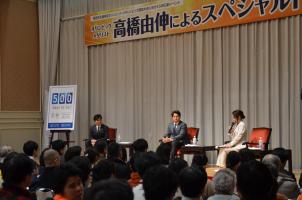 大勢の参加者が見ているなかステージ左側に男性と中央に高橋由伸氏が座り、右側に座っている女性がマイクを持ち話をしているトークショーの様子の写真