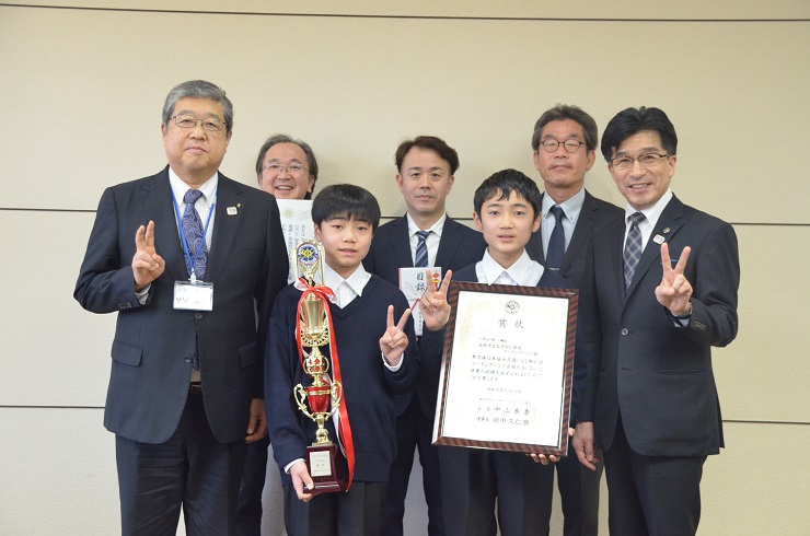 トロフィーと賞状を持った男子児童2名が真ん中でピースをし、両隣りの市長と男性もピースをしており、後ろに男性3名が並んで記念撮影をしている写真