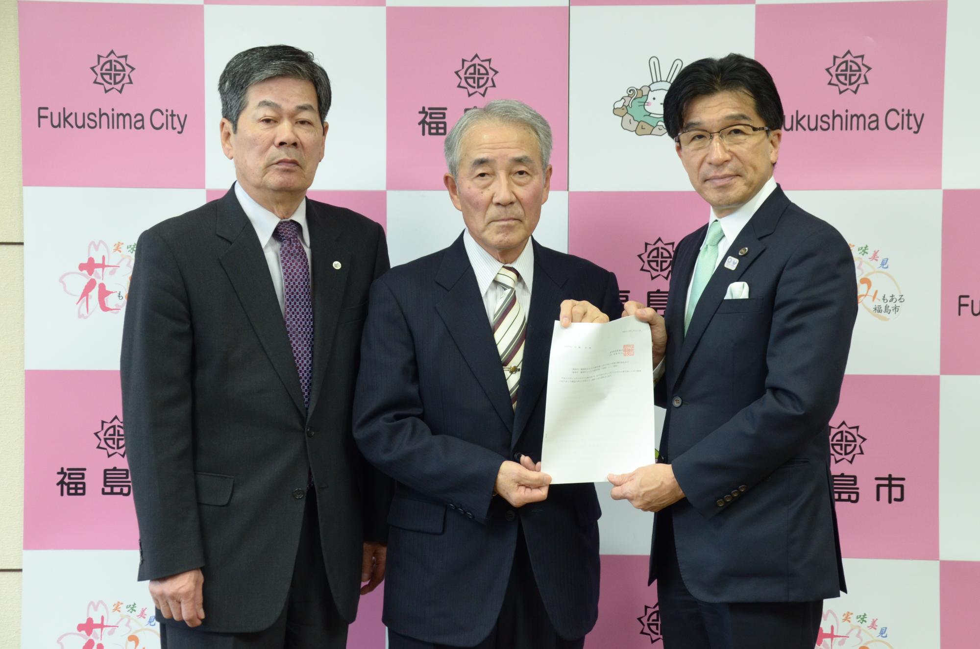福島市のバックパネル前で、市長へ答申書を手渡す高野会長と関係者の写真