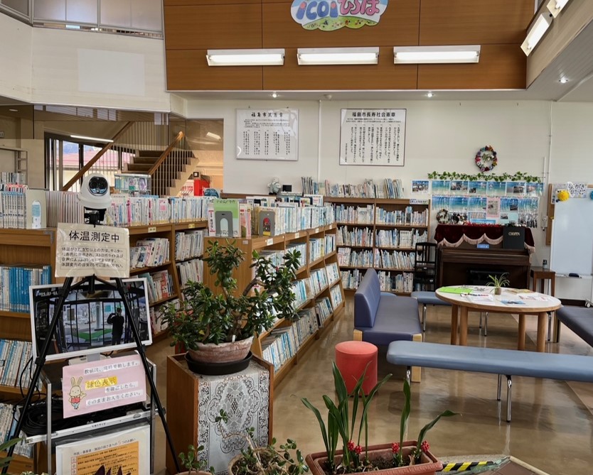 体温測定中と書かれた測定器の奥には丸いテーブルに長椅子がセットされ周りにはたくさんの本がならんだ本棚があるロビーの写真