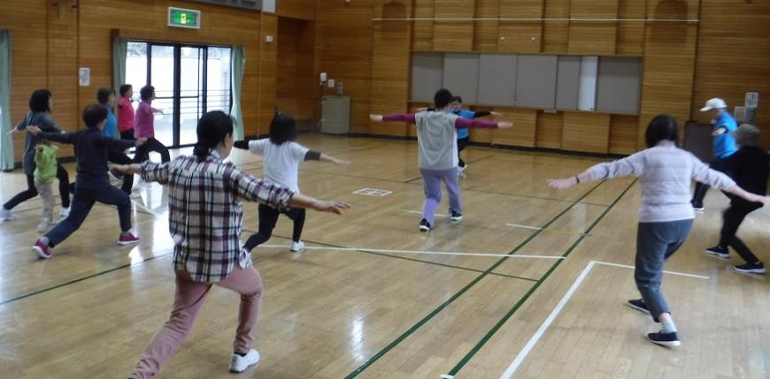 右足を下げて斜めにだし、左足は後ろに伸ばし両手を真横に伸ばして体操をしている参加者の人たちの写真