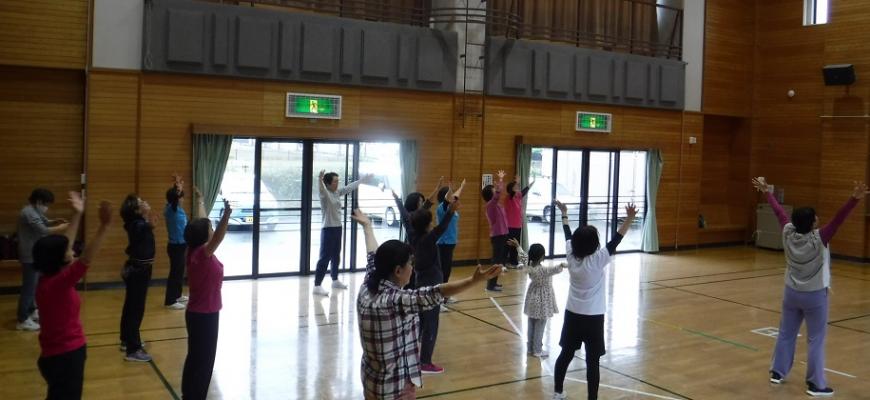参加者の人たちが両手を斜め上にあげて胸をそらして体操をしている体育館内での写真