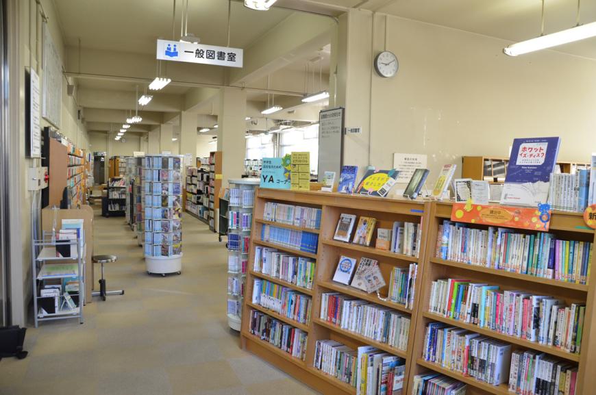 廊下にたくさんの本が入った本棚、天井から一般図書室の案内板が吊るされた奥にたくさんの本が展示されている市立図書館内の写真