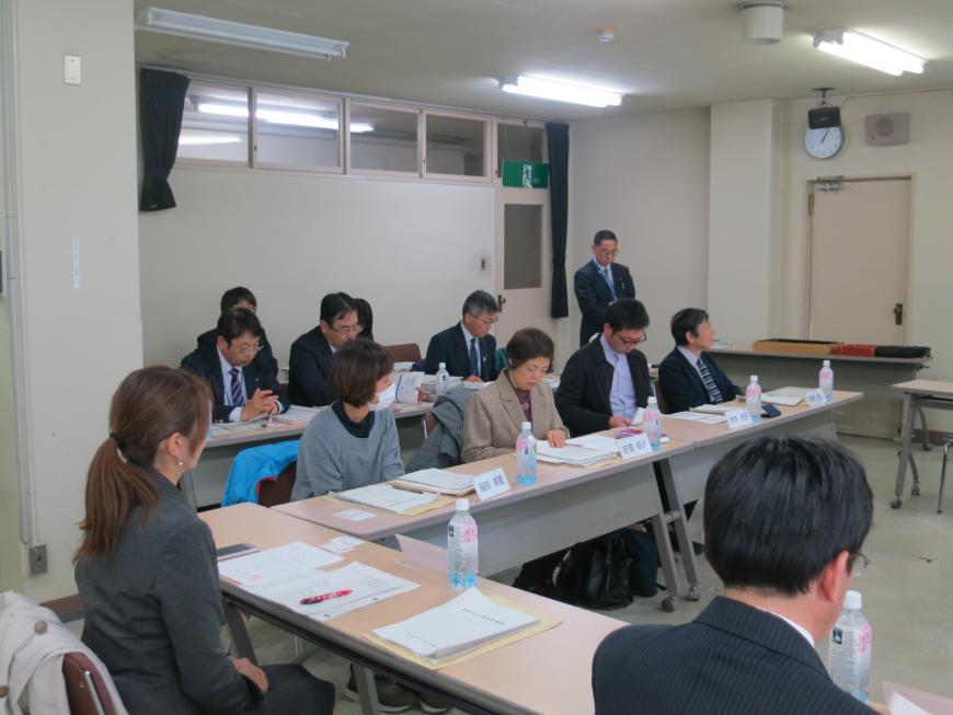 コの字型に配置された机に参加者の方が座り、その横で男性が立って話をしている写真