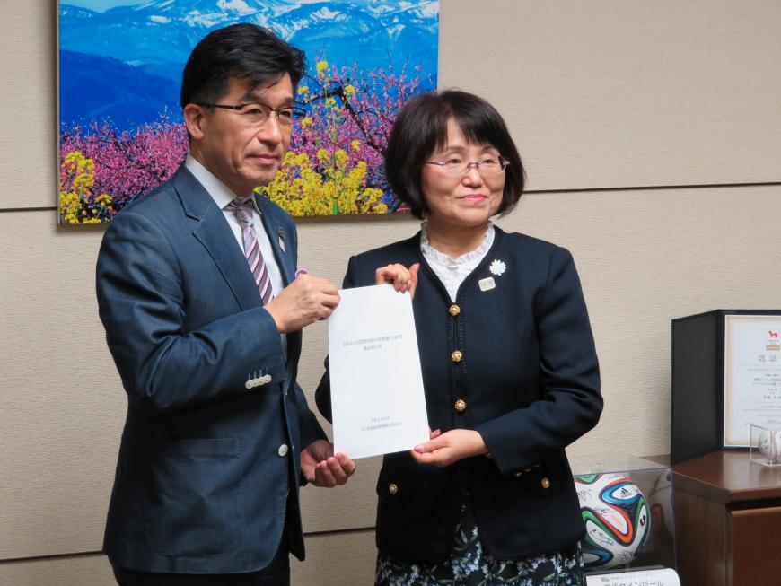 風景の写真が壁に飾られた部屋で、市長と女性で報告書を持って立っている写真