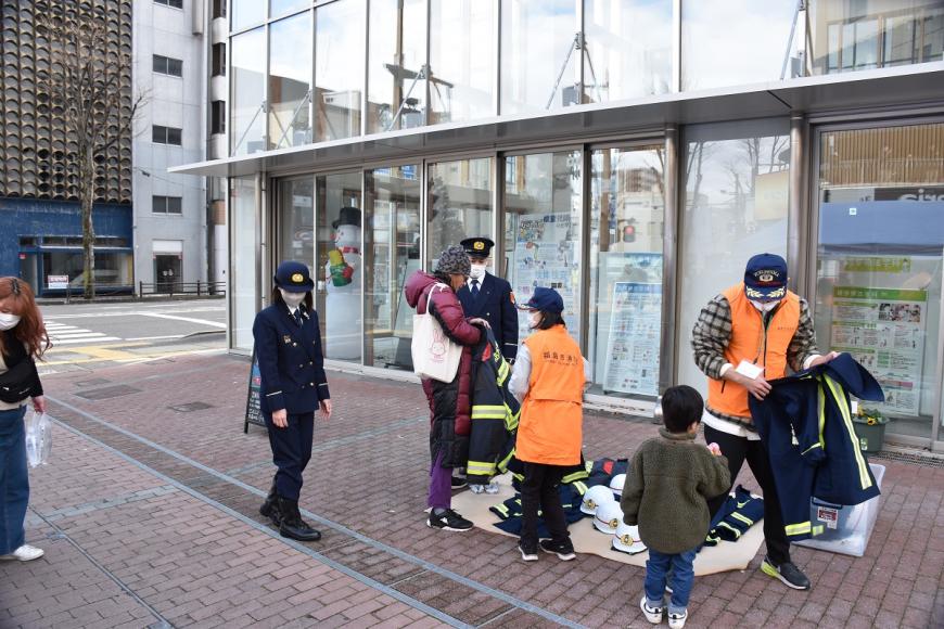 歩道の上に敷かれたシートの上に消防服やヘルメットが並べられ着装体験が行われている様子の写真