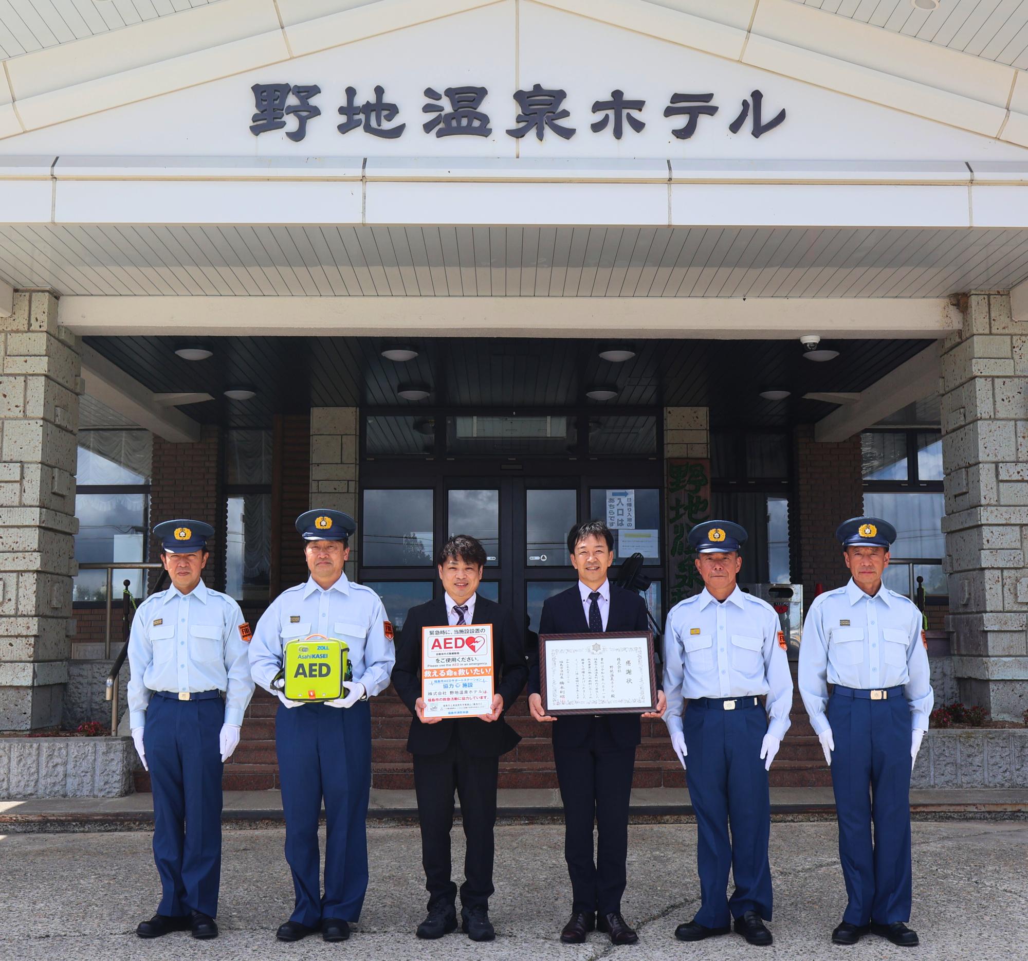 野地温泉ホテルエントランスにて、従業員代表2名が中央で感謝状とAEDサポートステーション登録掲示板を持ち、消防職員4名と記念撮影した写真