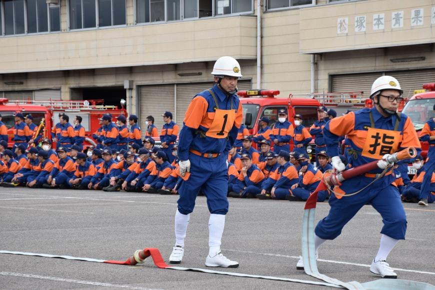 ヘルメットをかぶりオレンジ色のゼッケンをつけ、紺色の活動服を着た男性2名が、消防用の筒状の吸管の管を持ち上げて伸ばす操法技術を披露している写真