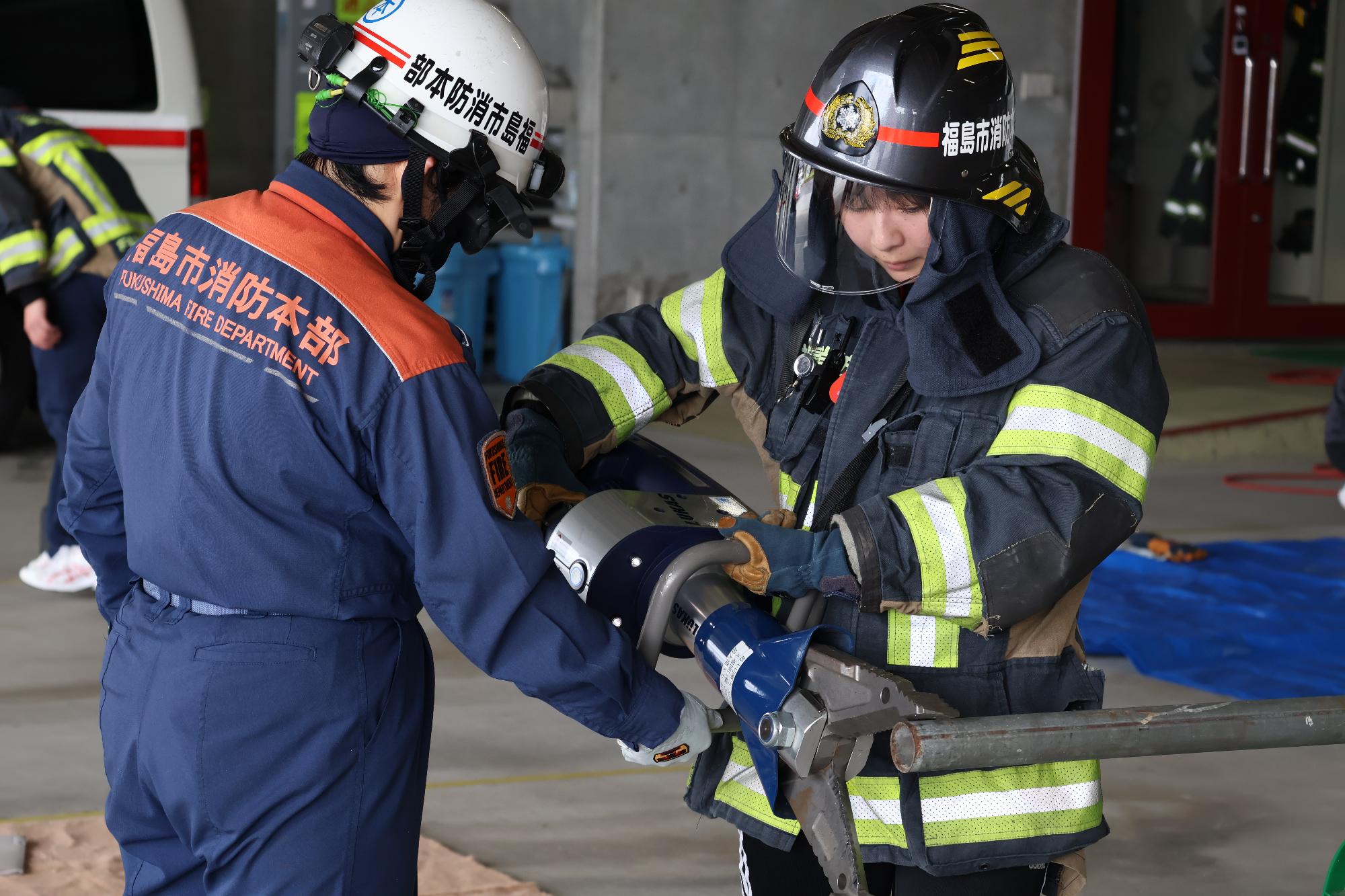 消防職員に見守られながら、防火服とヘルメットのシールドで防護した参加者が救助資機材で鉄管をつぶしている写真