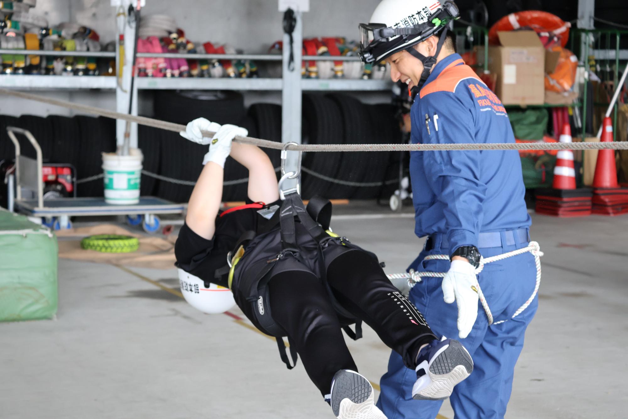 ヘルメットと手袋を着用した参加者が、消防職員に付き添われ空中に床と並行に張られたロープにぶら下がりながらロープ渡りをしている写真