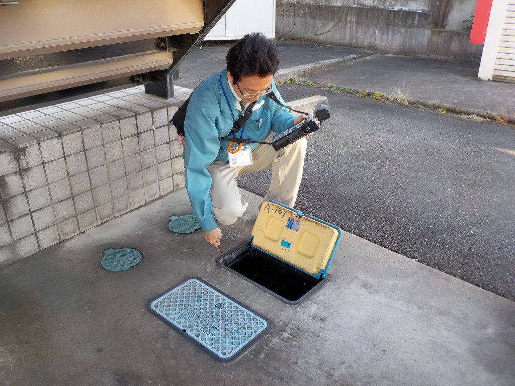 屋外で、作業服を着た男性が、開けられたマンホールの蓋のそばにしゃがみ込み、手に持った測定機器のようなものを操作しながら検針を行っている様子の写真