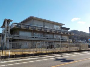 手前に黒いフェンスがあり、その奥に灰色のコンクリート造りで3階建ての水道料金お客さまセンターがある建物の外観写真