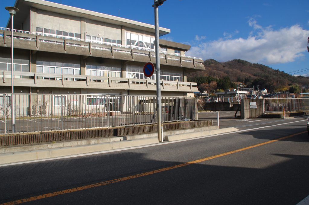 道路沿いに立つ3階建ての建物で、フェンスに囲まれ、建物前に駐車場がある水道料金お客さまセンターの外観写真